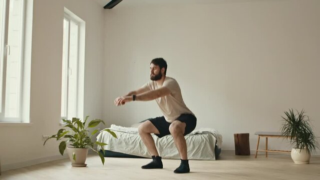 Home workout. Young bearded man doing squats exercises, sports training alone
