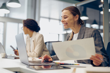 Smiling female accountant working with financial statement and use laptop while sitting in office 