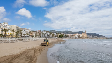 A bulldozer grooming a sandy beach in a scenic Mediterranean coastal town, with palm trees and mountains backdrop, ideal for travel and summer holiday themes