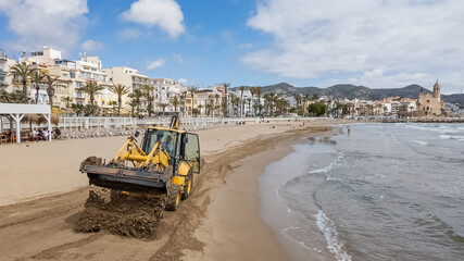A yellow bulldozer grooming sand on a Mediterranean beachfront promenade under a cloudy sky, prepping for tourist season  concepts: beach maintenance, travel destinations © fotoworld
