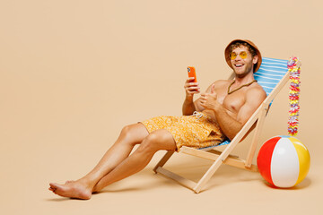 Full body young fun man wear yellow shorts swimsuit hat relax rest near hotel pool sit in deckchair hold in hand use mobile cell phone isolated on plain beige background. Summer sea sun tan concept.