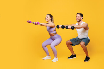 Full body side view young strong fitness trainer sporty two man woman wear blue clothes spend time in home gym do squats hold dumbbells isolated on plain yellow background. Workout sport fit concept.