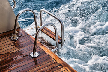 Back teak wooden deck and metal ladder of a of motor yacht, waves and sea splashes and foam.