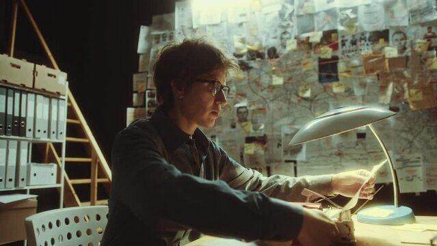 Female detective typing text on paper with typewriter and attaching it to evidence board, then examining clues and thinking about crime investigation during night in the office