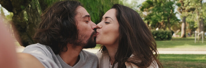 Happy couple kissing and taking a selfie sitting on blanket in park, panorama