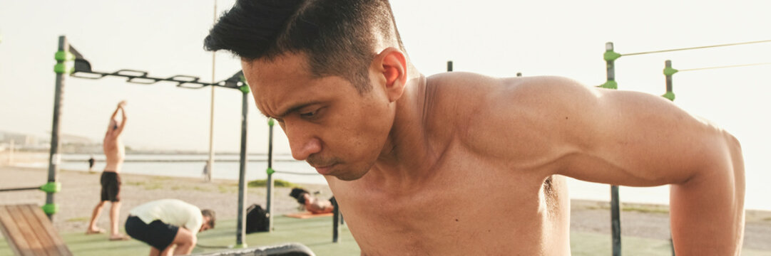 Guy, the athlete does push-ups on the parallel bars on the outdoor sports ground on a summer day, panorama
