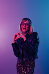 Lovely beautiful blonde woman in leather jacket sparkly dress trendy sunglasses show heart with fingers posing isolated in blue pink color light studio background. Neon party concept. Copy space
