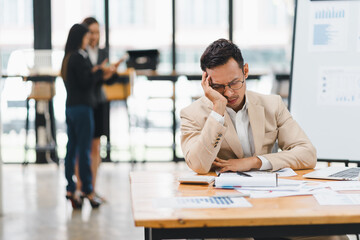 Businessman feeling stressed while sitting at desk cluttered with documents and a laptop in a modern office.
