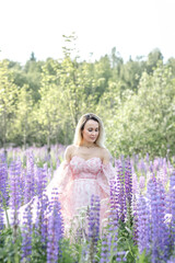 Beautiful girl with blond hair and a long pink dress in the summer in a field of blooming purple lupine flowers with a bouquet in her hands