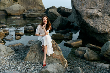 Serene woman in white dress sitting on ocean rock gazing at large boulders