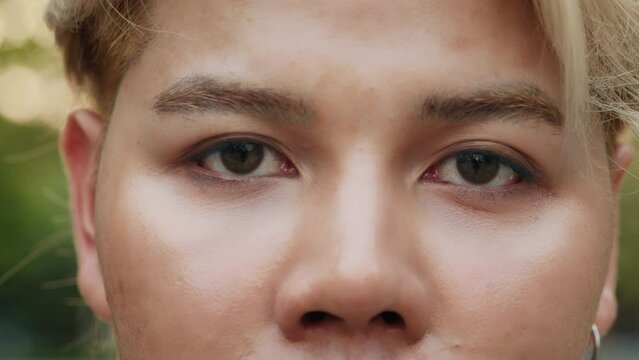 Young Asian man with bleached hair and chocolate eyes outdoors closeup. Handsome male model with languid look in green park. Beauty and fashion industry attractive guy job