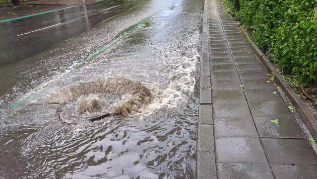 Water lifting an iron sewage manhole cover on the road, strong water pressure flowing onto the roadway. Impromptu flooding after heavy rain, drainage systems overwhelmed by the force of nature.