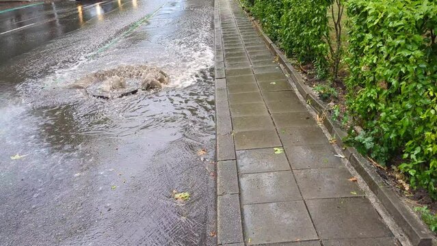 Water lifting an iron sewage manhole cover on the road, strong water pressure flowing onto the roadway. Impromptu flooding after heavy rain, drainage systems overwhelmed by the force of nature.