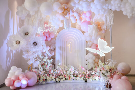 Birthday party for 1 year old girl on a background photo wall. Arch decoration pink, white balloons, text Happy Birthday, flowers, paper decor butterfly. Children's photo zone.