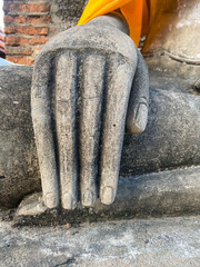 Close-up of weathered stone Buddha statue's hand on lap, in vibrant yellow robe, against brick wall