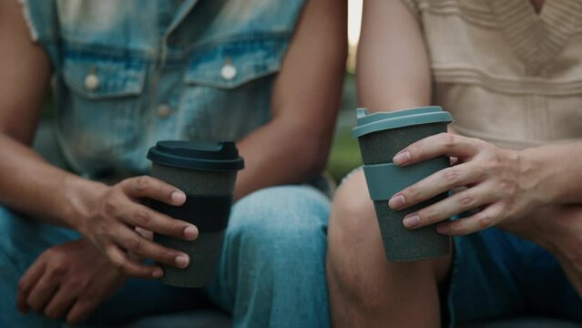 Gay couple holds coffee cups sitting nearby in urban park closeup. Young men from LGBTQI community spend time together on date. Celebrating pride month for gay and queer people support