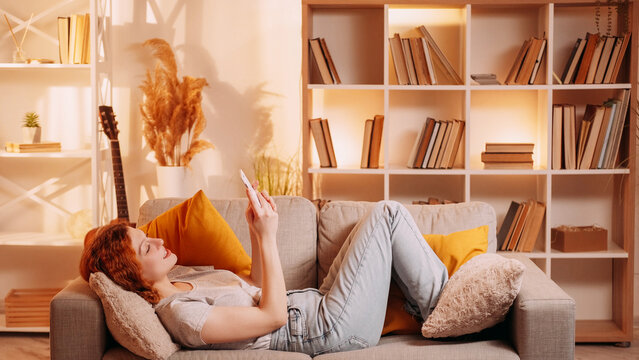 Video call. Mobile leisure. Online communication. Relaxed girl speaking on phone camera resting on couch at warm home interior with free space.