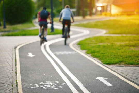 Curvy bike lane with markings and men cycling in the background, recreational and fitness benefits of cycling. Bicycle lane in park. Bicyclists riding. Selective focus. - Powered by Adobe