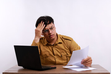 Stressed male teacher in civil servant uniform with paper and laptop on the desk holding a paper