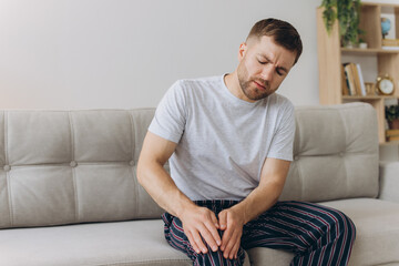 Unhappy man sitting on sofa at home and touching hurt knee with grimace of pain and suffering....