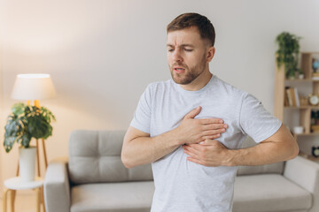 A young man is standing at home and holding his chest. Feels severe pain, shortness of breath,...