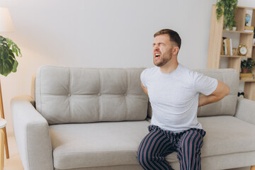 Fototapeta premium Tired bearded man suffering from back pain sitting on couch in living room at home. Male massages painful lower back muscles. Caucasian guy exhausted overworked in apartment.
