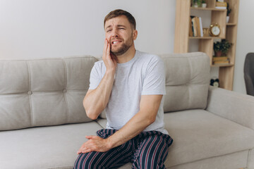 Dental health problems concept. Man suffering from strong toothache. Upset male patient with inflamed nerve, tooth abscess, or teeth sensitivity sitting on sofa and touching cheek with grimace of pain