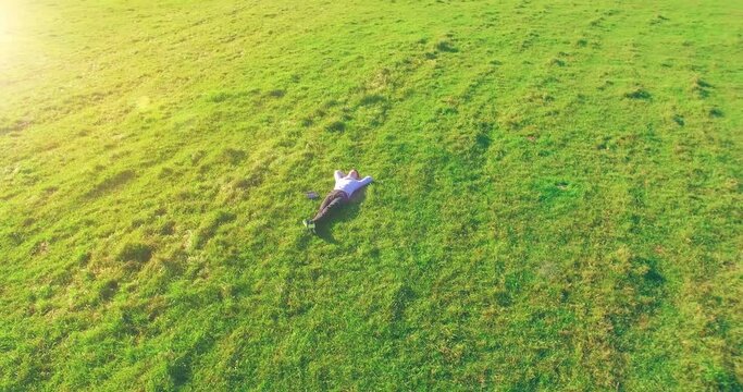 Aerial view point. Low orbital flight around man lied on green grass with notebook (pad) at yellow rural field. Casual weared. Sun rays. Radial movement.