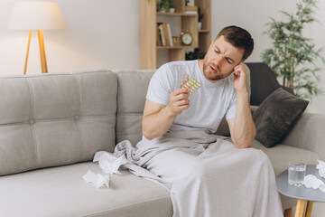 Sick man suffering from fever and headache holds blisters with pills while sitting on the sofa....
