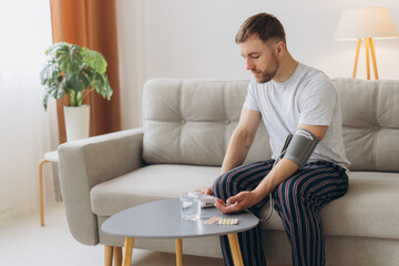 Young caucasian man sitting on couch at home, checking blood pressure. Bearded guy in pajamas using...