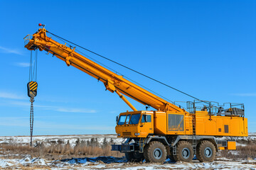 Yellow Mobile Crane on Snowy Field with Extended Arm and Hook Against Clear Blue Sky