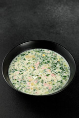 Cold soup with fresh cucumbers, sausage, broth and sour cream in a black plate on a black background. A traditional dish is okroshka