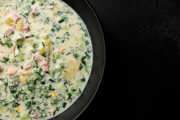 Cold soup with fresh cucumbers, sausage, broth and sour cream in a black plate on a black background. A traditional dish is okroshka
