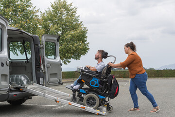 Female assistant transferring a man with disability into the vehicle by pushing his wheelchair up...