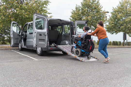 Female assistant transferring a man with disability into the vehicle by pushing his wheelchair up the accessible car ramp.