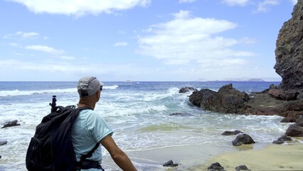 The man in the hat looks at the sea from the sandy beach and lava rocks. The man is a young adult and is photographed from behind. The ocean is green and turquoise in color. Summer travel concept