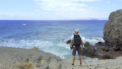 A male hiker contemplates the ocean from the lava rock shore. The man is a young adult and Hispanic. The boy is photographed standing and from behind. Freedom and summer travel concept