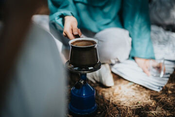 Close-up of a person brewing coffee on a portable stove in the forest, capturing an outdoor camping experience and relaxation.
