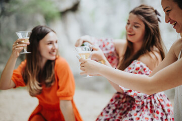 Three young women enjoying a sunny picnic, smiling and toasting with champagne glasses outdoors, capturing a carefree and joyful atmosphere.