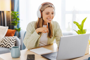 Young woman wearing headphones taking notes while studying online at home