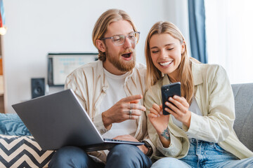 Young couple sitting on sofa at home using laptop and smartphone browsing social media having fun