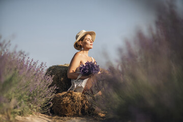 woman sitting in a field of lavender and wearing a straw hat. She is smiling and holding a bouquet of flowers. Scene is peaceful and serene, as the woman is surrounded by the beauty of nature