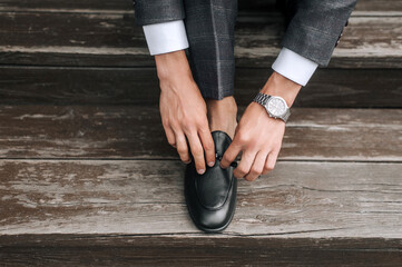 Stylish fashionable male businessman, young groom in a gray suit fastens black leather shiny shoes...