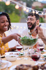 Vertical. Smiling African American woman sitting at table passing salad to friends at meal event...