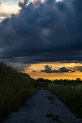 sole che tramonta e colora il cielo di arancione mentre delle nuvole molto scure di colore blu di un temporale avanzano sopra un territorio di campagna, visto da una strada sterrata