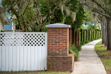 The exterior corner fence of a neighborhood is made of wood boards and white lattice. The yard is a divider of red brick with trees, foliage, and plants. A walking path skirts the fence of trees