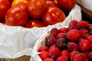 Two small baskets with white plastic bags covering the bowls. One has small orange tomatoes the other has dwarf crab apples, prunus avium. The organic fruit is for sale in bulk at a farmer's market.