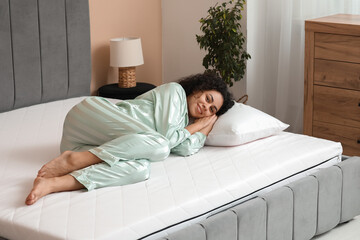 Young African-American woman with pillow lying on soft mattress in bedroom