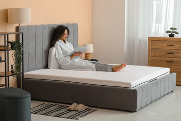 Young African-American woman reading magazine on soft mattress in bedroom