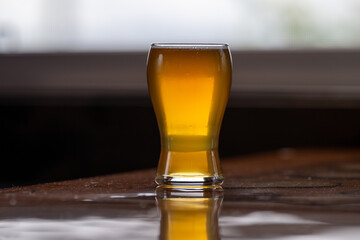 A small curved tulip shaped sample glass of pale ale beer with froth on the top. The liquid alcohol has a vibrant yellow tint. The glass is on a wooden table with a glossy top. The ale is orange color